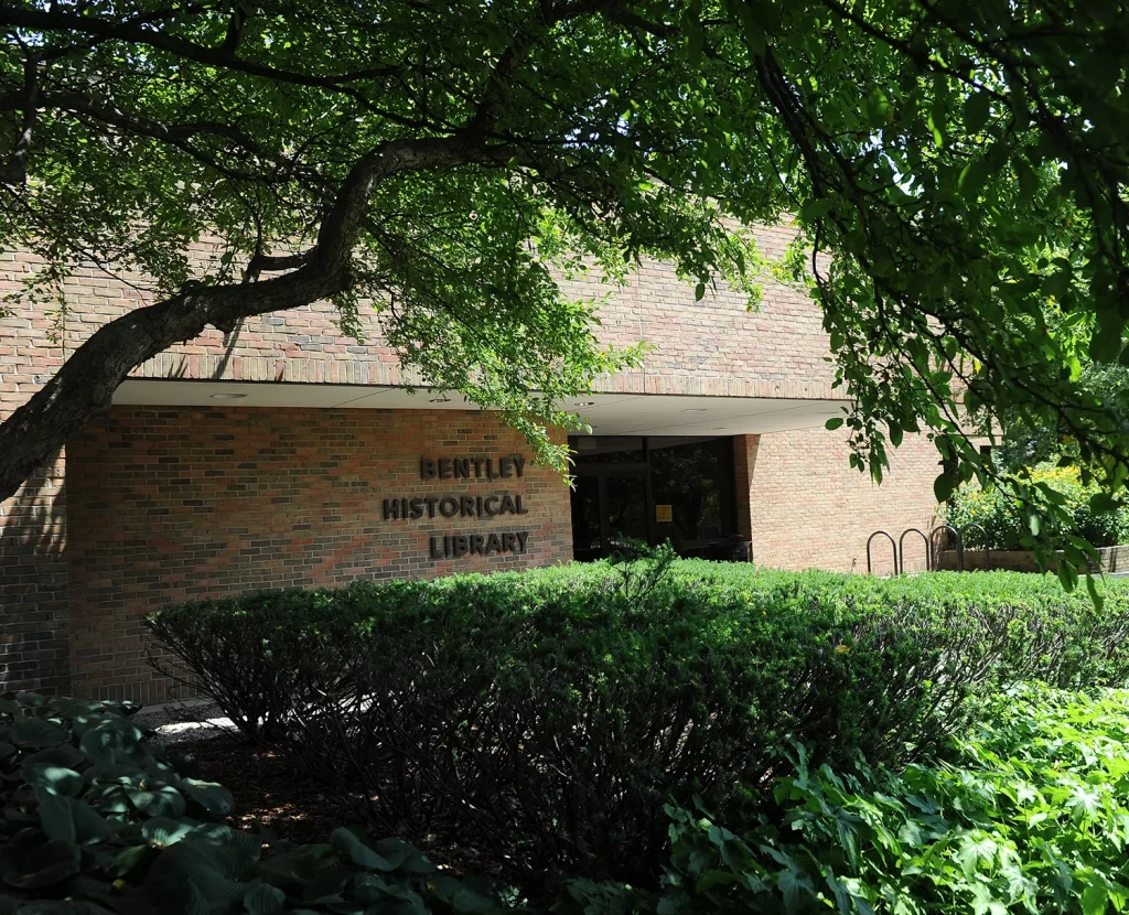 Green bushes and branches frame a picture of the Bentley Historical Library's brown brick exterior. The library's sign and name is visible in the photo.