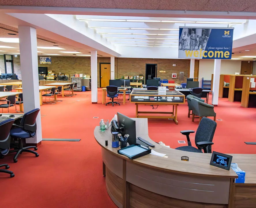 A large room with red carpet contains many desks and tables. The welcome desk of the Bentley's reading room is visible. A banner hanging above it says "welcome."
