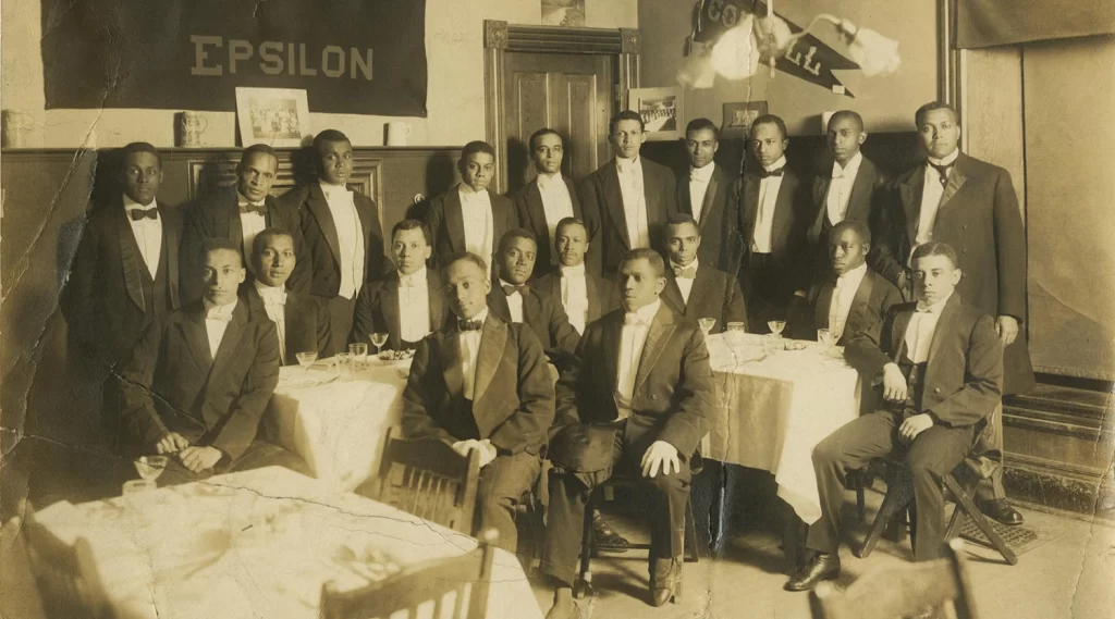 African American members of the Alpha Phi Alpha fraternity sit on either side of a long white table wearing bowties and formal suits.