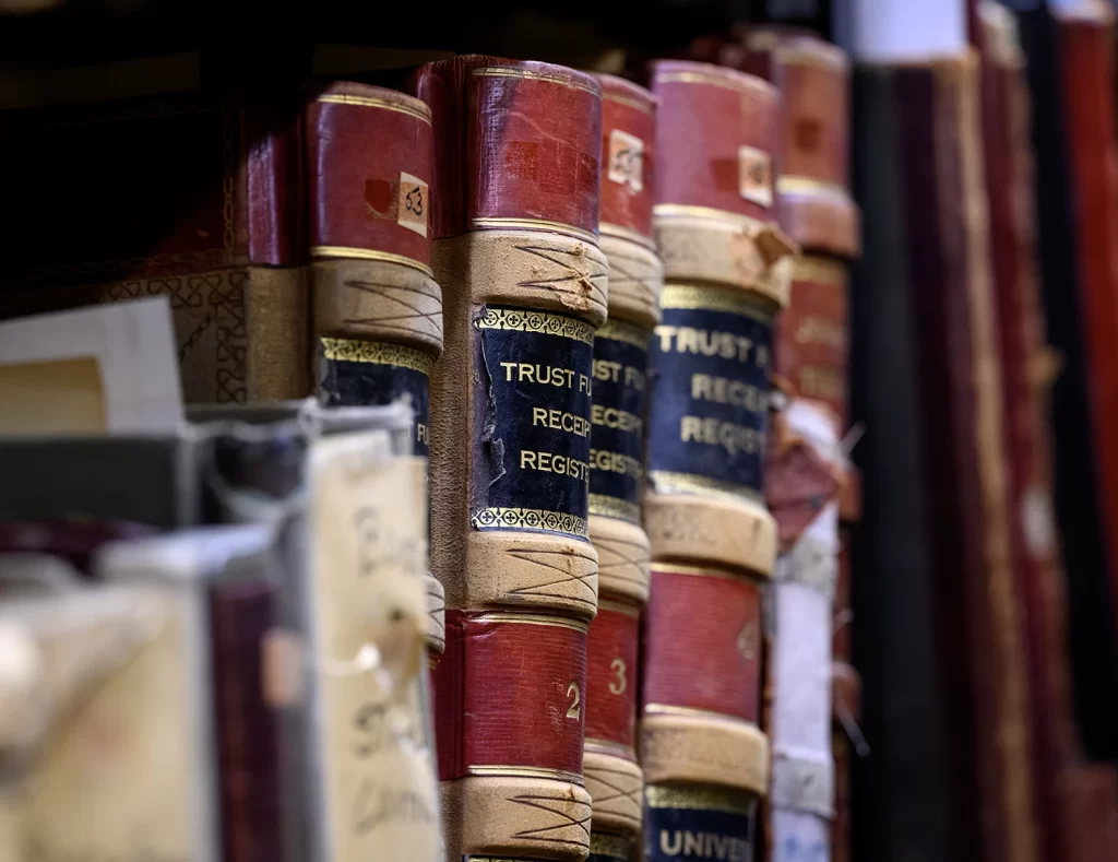 A side view of a row of historical books on a shelf inside the archives at the Bentley Historical Library, featuring book spines bound in red leather and tan suede with gilded lettering, with visible words on the spine reading: Trust, Receipt, Register.