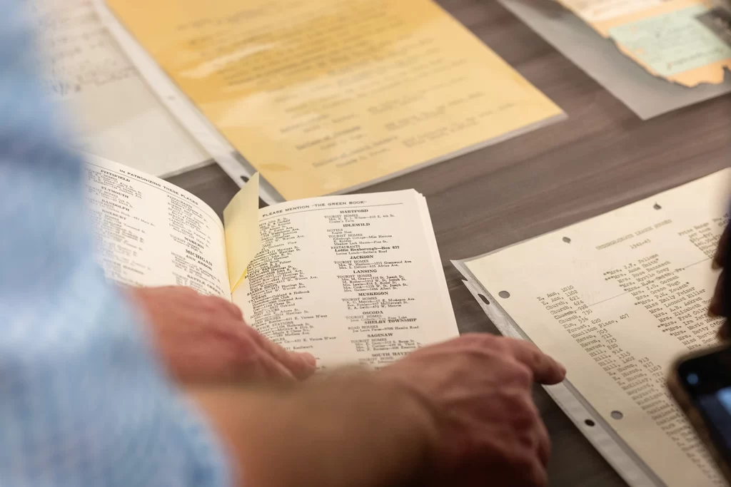 Papers on a desk with a set of hands holding a book open and pointing to figures.
