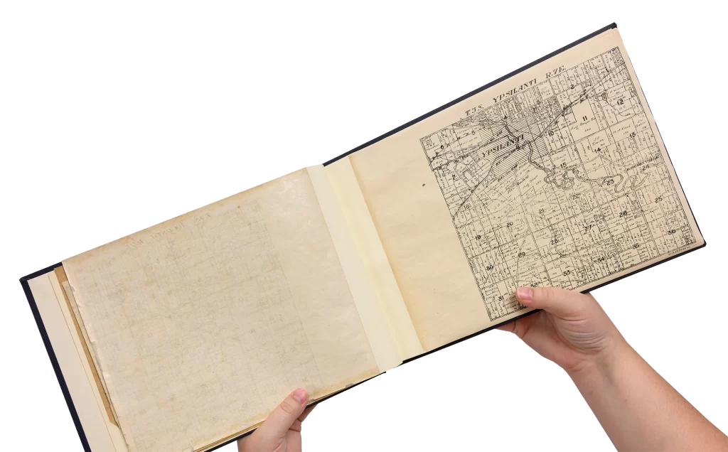 Two hands holding an open Plat Book full of maps of land ownership in Michigan areas, showcasing a map of Ypsilanti.