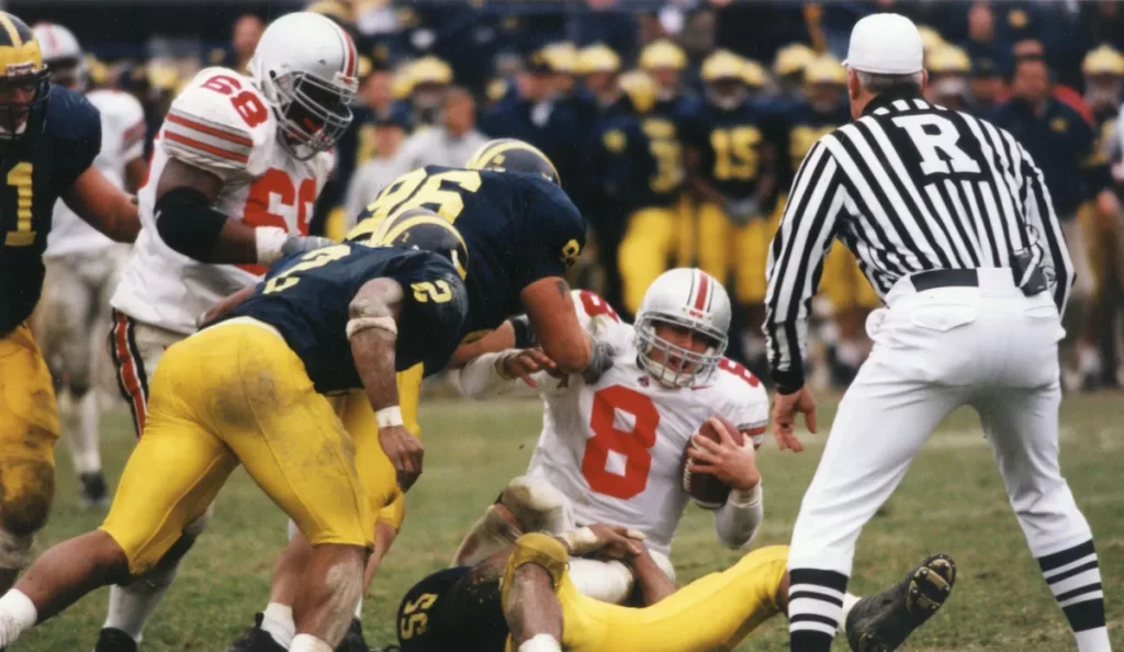 A football player wearing the number 8 jersey is down on the field holding the football, while University of Michigan players to the left run toward him. A referee in a black and white shirt stands to the right.