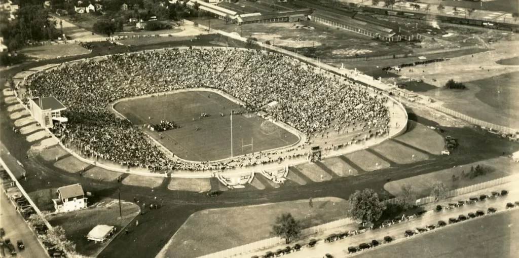 A black-and-white aerial photo over Michigan Stadium from 1927 on the stadium's opening day. Fans are visible in the stands, with a cluster of people on the field. Houses, fields, and roads surround the stadium.