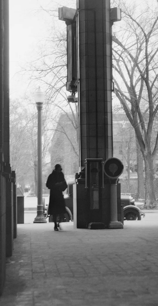 A black and white photo showing a woman wearing a long coat and hat. Behind her is an old car and campus buildings in the snow.
