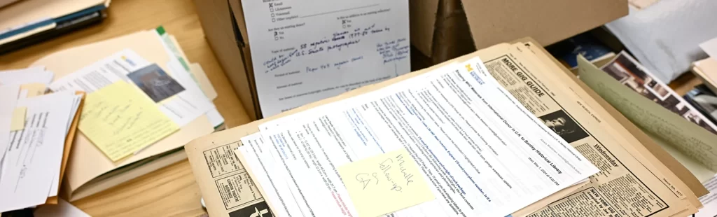 Papers and notes and photos arranged in piles on a desk, awaiting processing at the Bentley Historical Library.