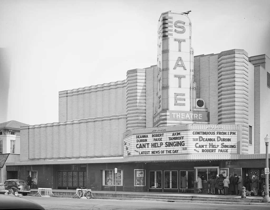 A black and white photo showing a large "State Theater" sign and a movie marquee with showtimes and movie titles. There are people lined up on the sidewalk outside to purchase tickets.