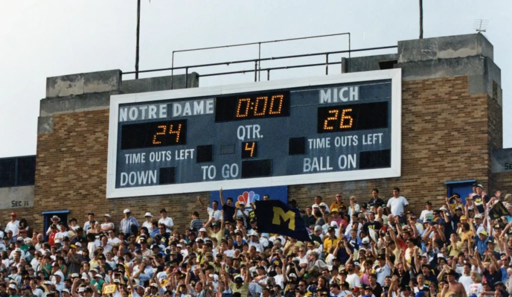 A large football scoreboard on a brick wall shows a score of Notre Dame, 24; Michigan, 26. Many fans are packed into stadium seats in front of the scoreboard, several of whom have their fists raised in the air.