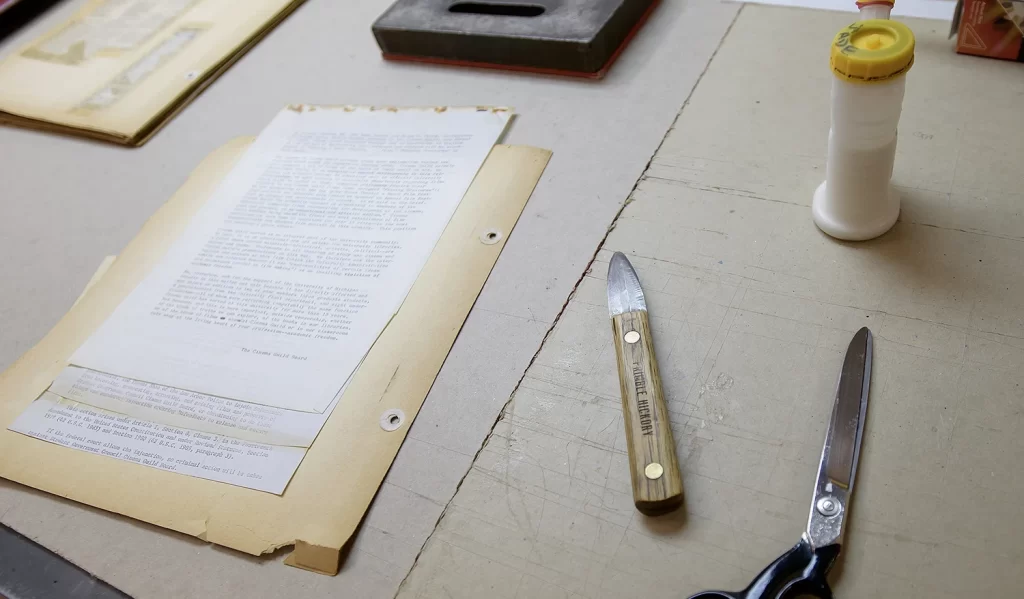 A historical page from the archives being worked on in the Conservation Lab at the Bentley Historical Library, including conservation tools like thread, scissors, and a metal scraper with a wooden handle, and a typed page of writing backed by a larger yellowing piece of paper with two punched holes on one side.