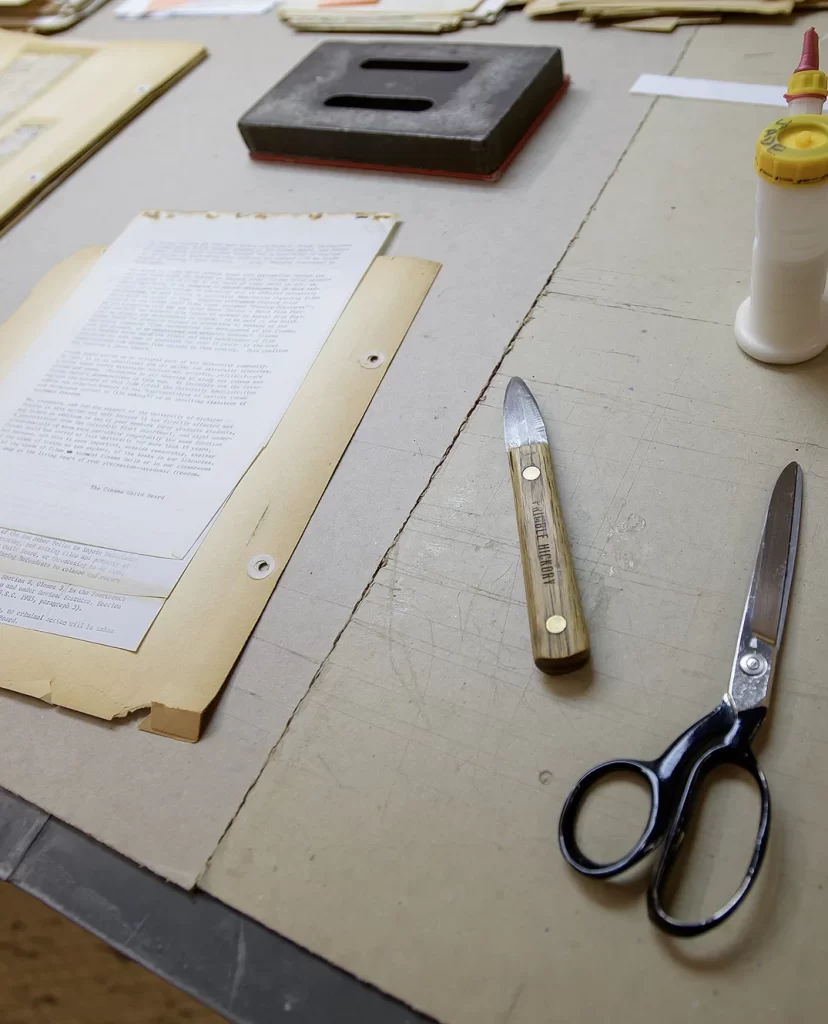 A historical page from the archives being worked on in the Conservation Lab at the Bentley Historical Library, including conservation tools like thread, scissors, and a metal scraper with a wooden handle, and a typed page of writing backed by a larger yellowing piece of paper with two punched holes on one side.