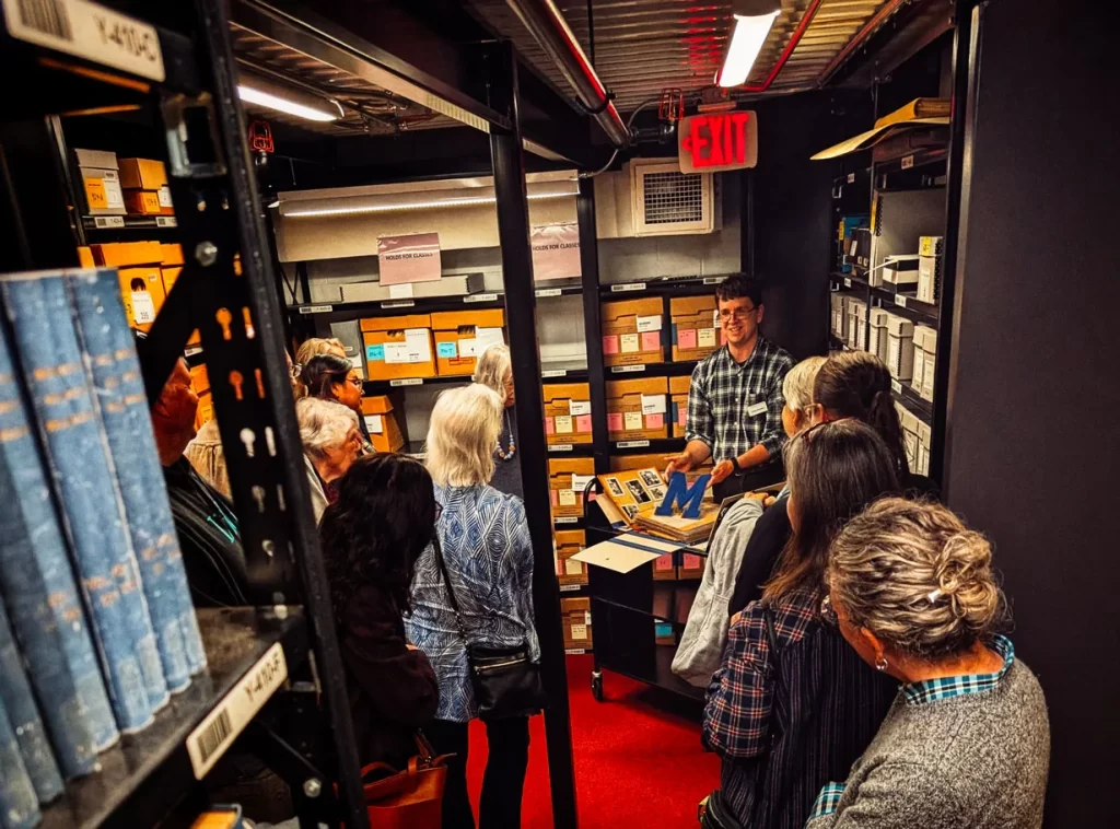 A group of people surrounds a man in the Bentley Historical Library's stacks giving a tour and pointing to a book.