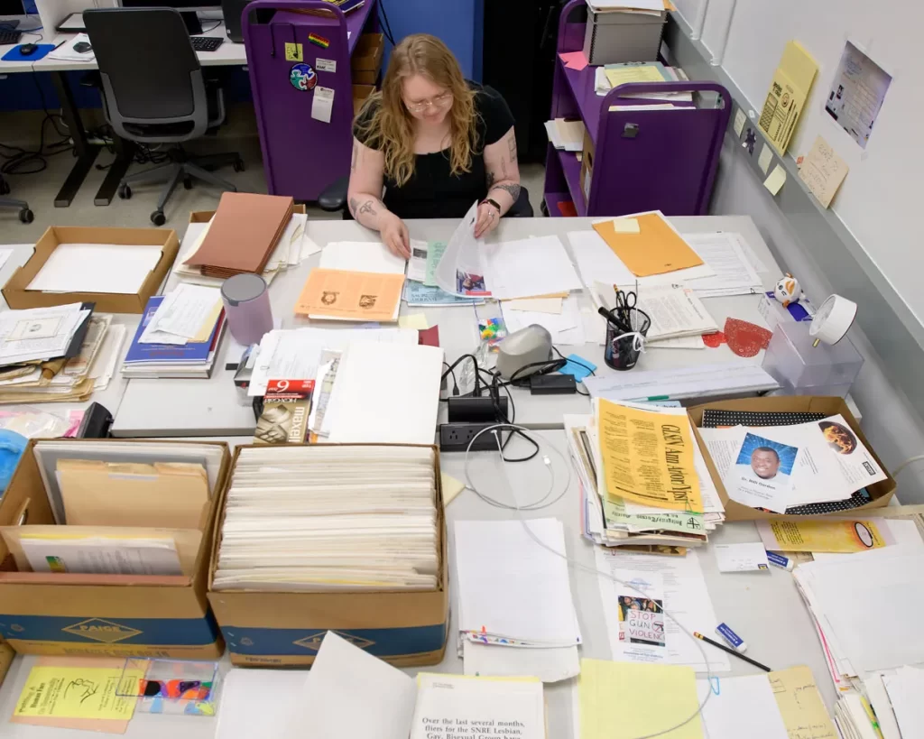 A woman sits at a table filled with papers and boxes, organizing the content as a processing archivist.