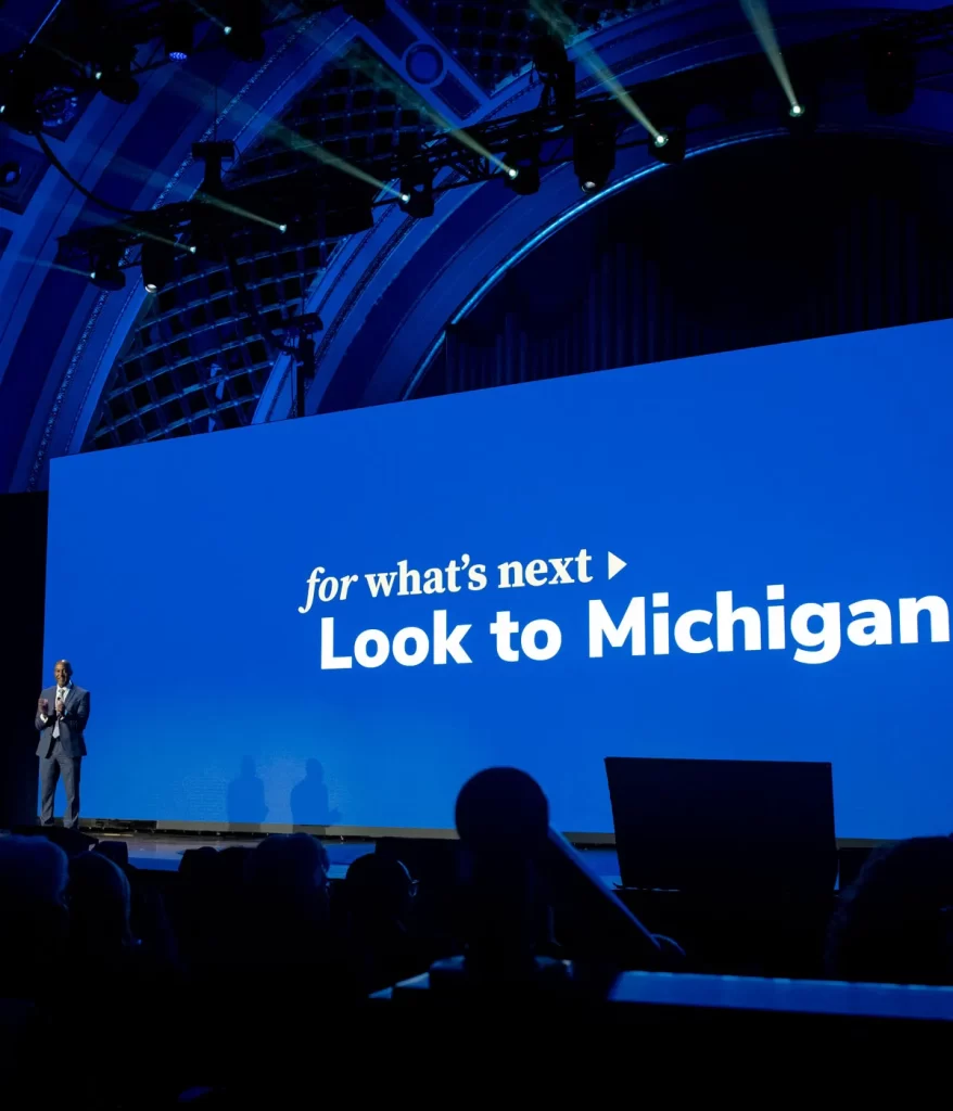 A blue screen projects white letters that say "For what's next, look to Michigan." The screen is on a stage. To the left of the stage, a man stands and applauds.