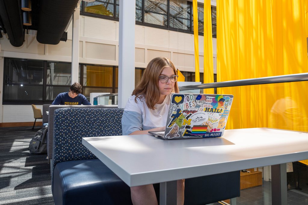 A woman wearing a white shirt sits at a table with a laptop open in front of her.