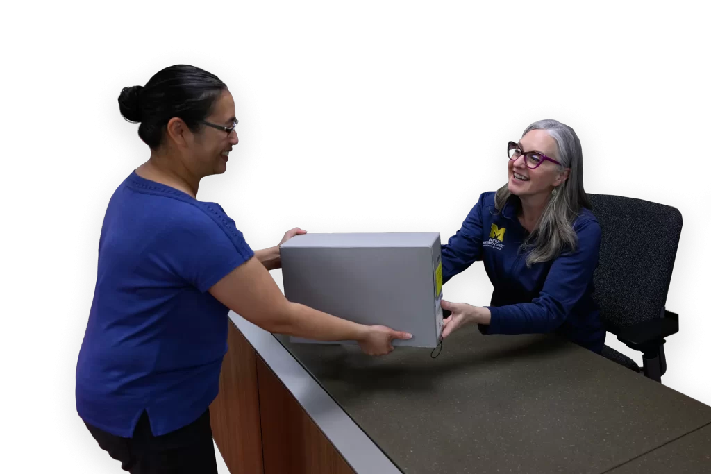 A woman in a blue shirt hands a gray box across a desk to another woman, also wearing blue.